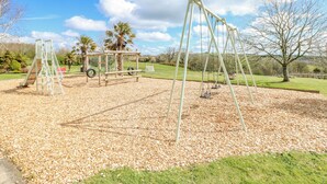 Une aire de jeux avec des balançoires et un toboggan, entourée d’une pelouse et d’arbres.