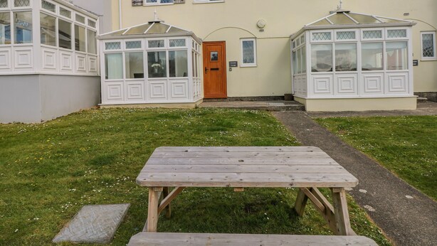 A two-story house with a front door, two bay windows, and a wooden bench in the front yard. - Grooms Cottage (Tenby)