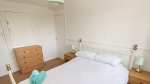 A neatly made bed with white linens, a wooden dresser, two bedside tables with lamps, and a door in the background. - Grooms Cottage (Tenby)