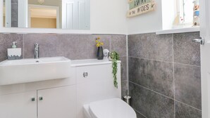 A modern bathroom with a white sink, mirror, and toilet. A decorative sign is on the wall, and a plant is on the counter. - Grooms Cottage (Tenby)