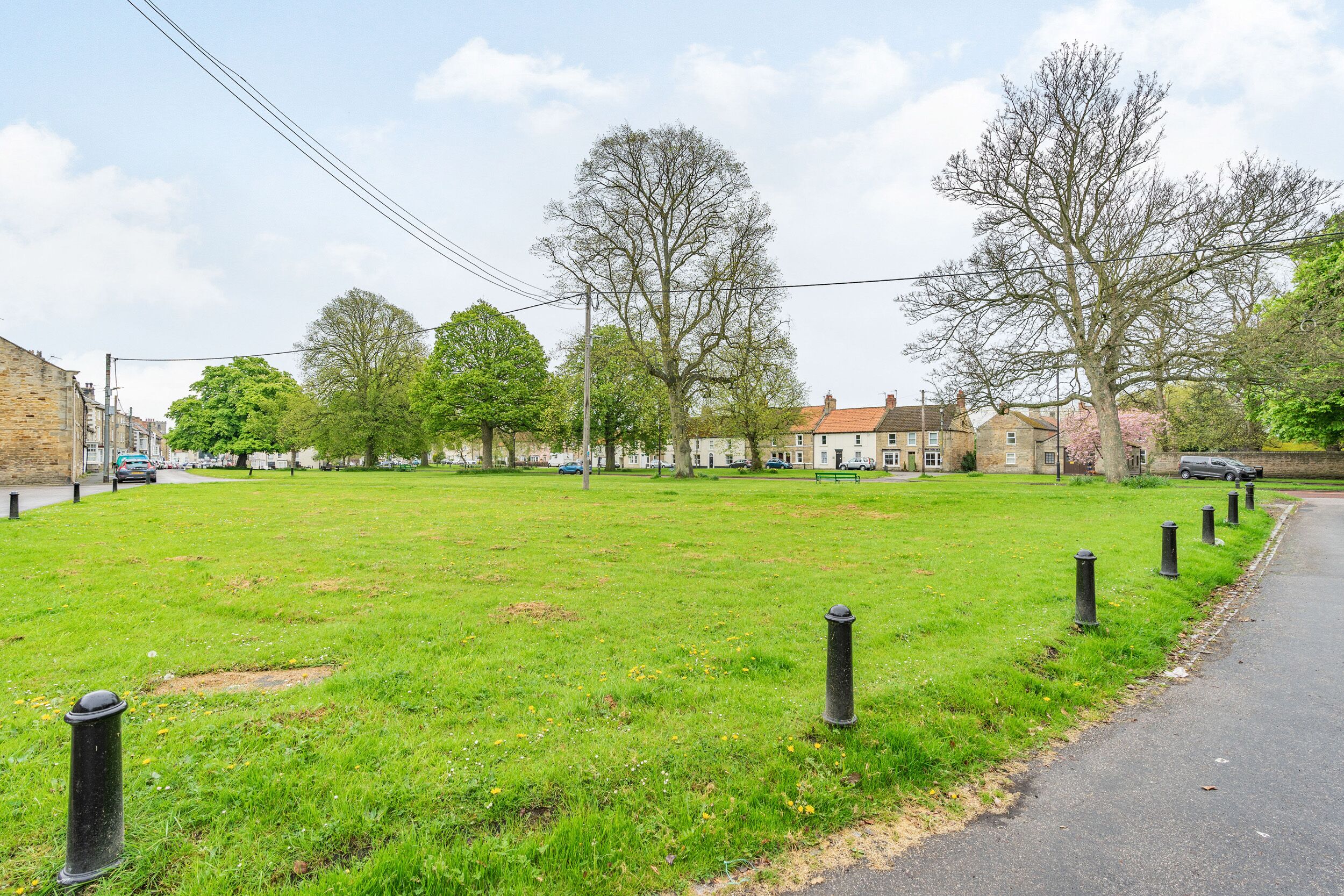 A grassy area with a paved path, trees, and residential buildings in the background.