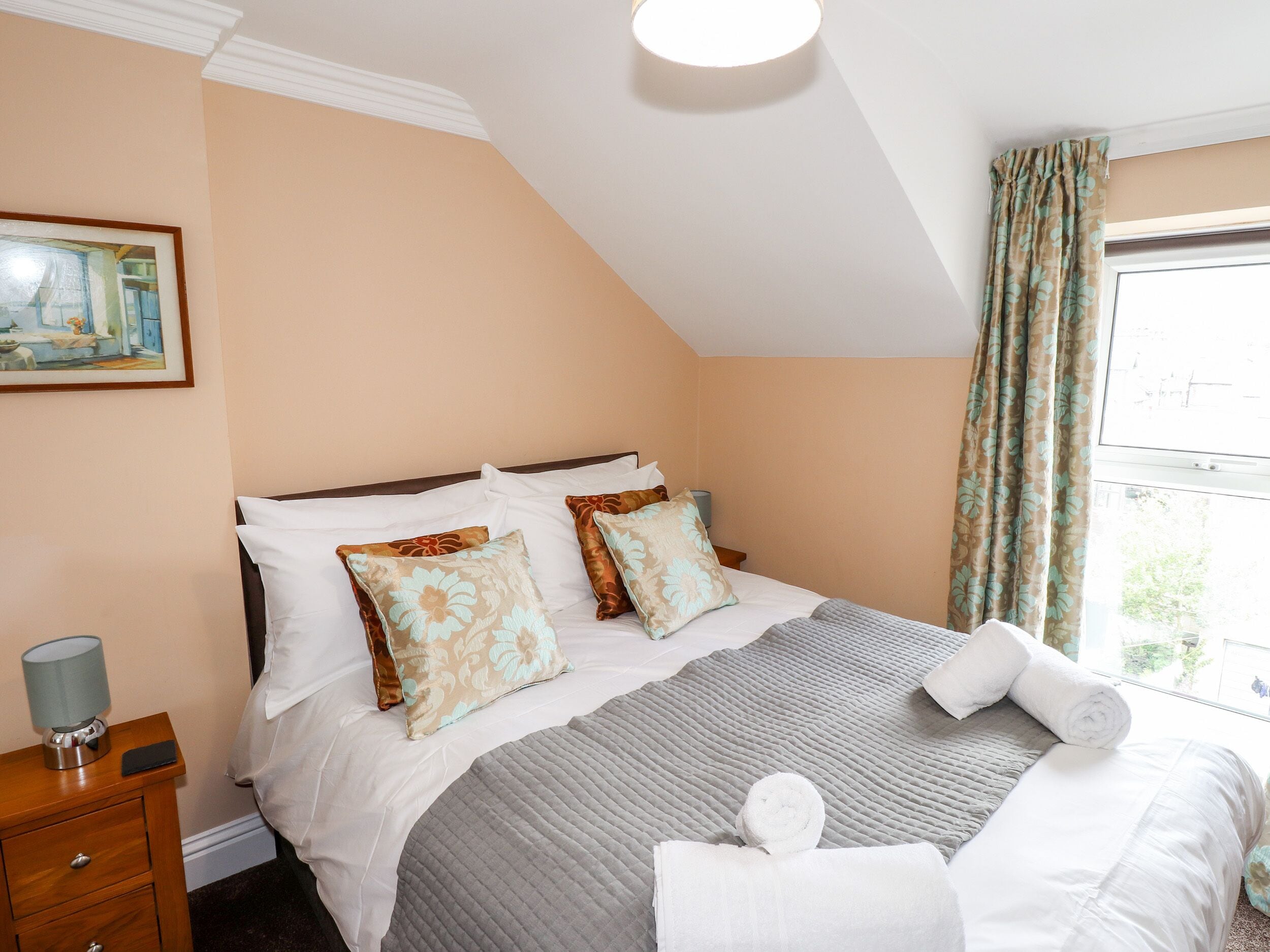 A neatly made bed with floral pillows, a bedside lamp, and a window with curtains.
