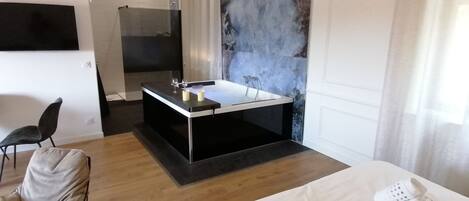 A modern hotel room with a large bed, a sitting area with a chair, a bathtub, and a shower.