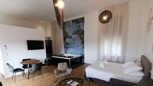 A modern hotel room with a bed, a sofa, a round coffee table, a desk with a chair, and a TV mounted on the wall. - Luxury Suite with Spa - La Nébuleuse (Vendres)