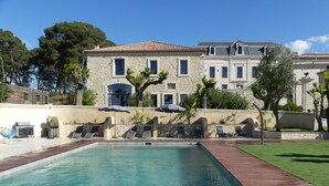 A swimming pool with a wooden deck, surrounded by lounge chairs and umbrellas, with a stone building in the background. - Luxury Suite with Spa - La Nébuleuse (Vendres)