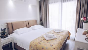 A hotel room with a large bed, a bedside table with a plant, a desk with a vase of flowers, and a window with curtains.