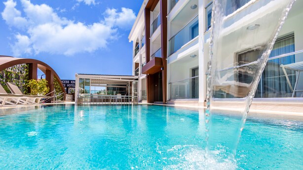 A clear blue pool with a water feature, surrounded by a modern building with multiple balconies and a glass facade. - Princess Kinli Suites Hotel (Marmaris)