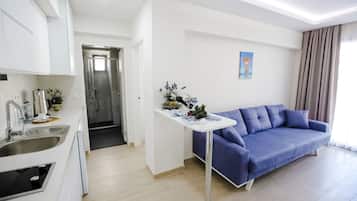 A modern living room with a blue sofa, a small white table with a vase, and a kitchen area with white cabinets and stainless steel appliances.