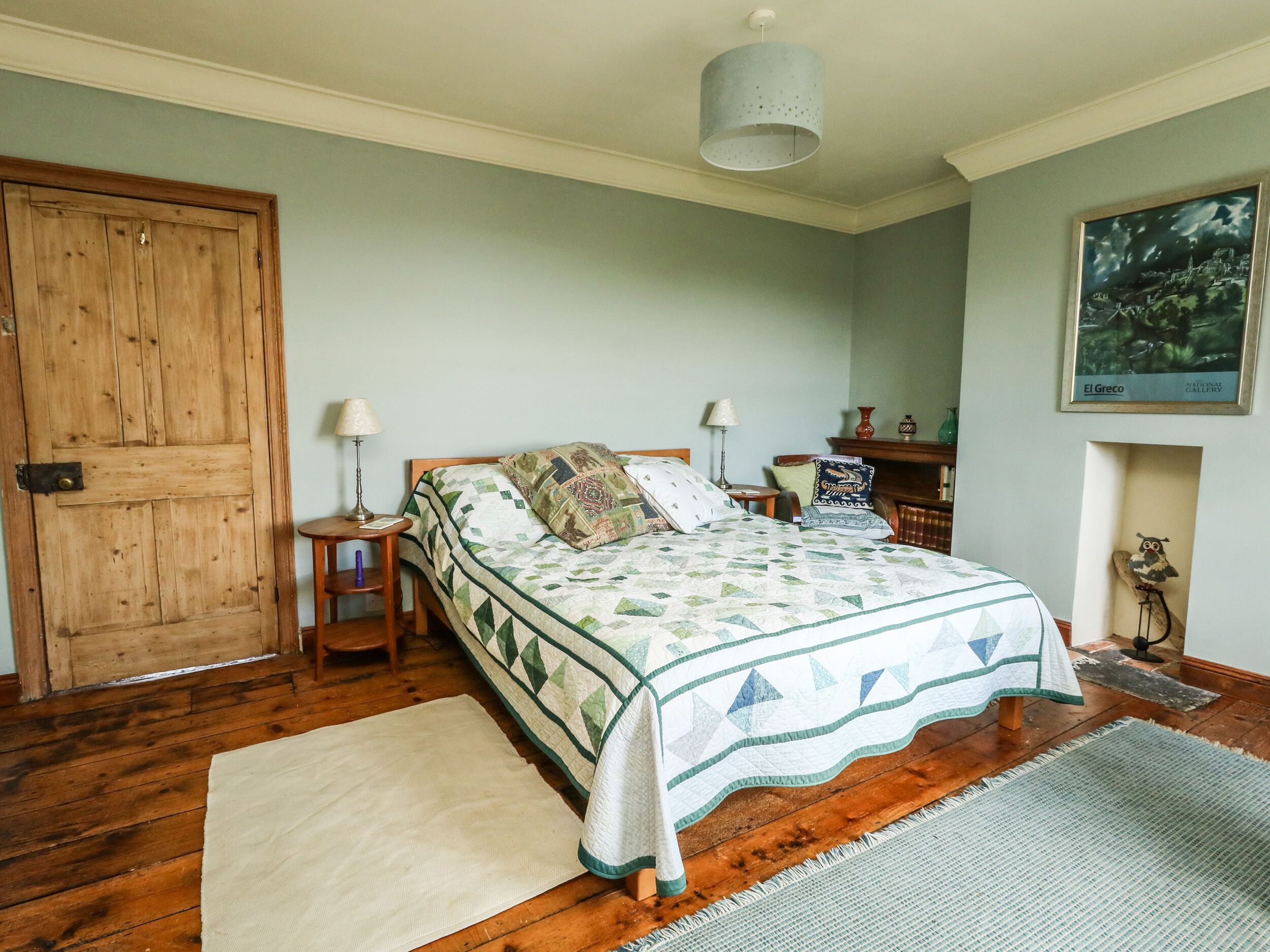 A bedroom with a wooden door, a bed with a quilt, two bedside tables with lamps, a rug, and a framed picture on the wall.