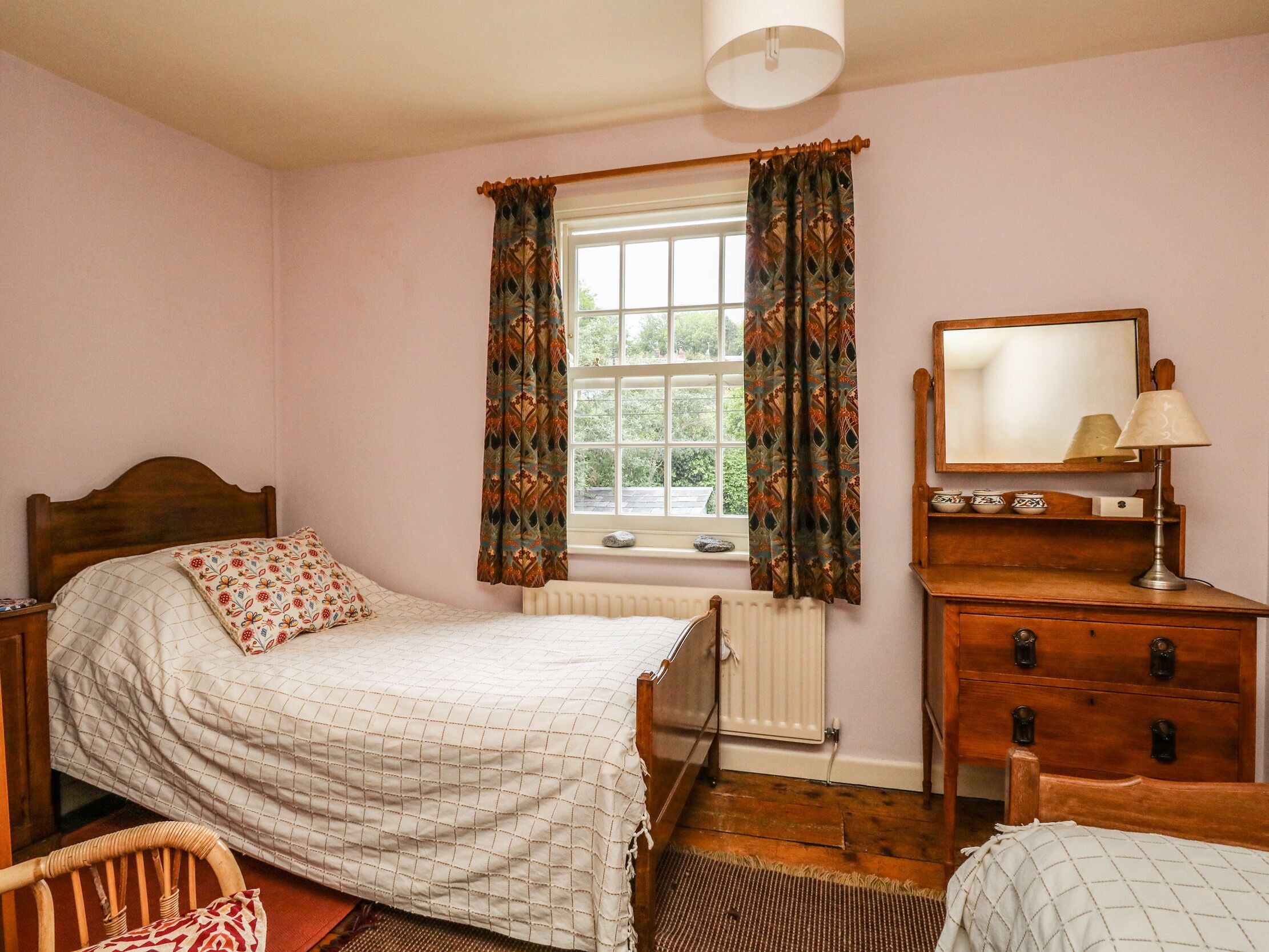 A bedroom with a bed, a dresser, a chair, a window with curtains, a radiator, and a mirror.