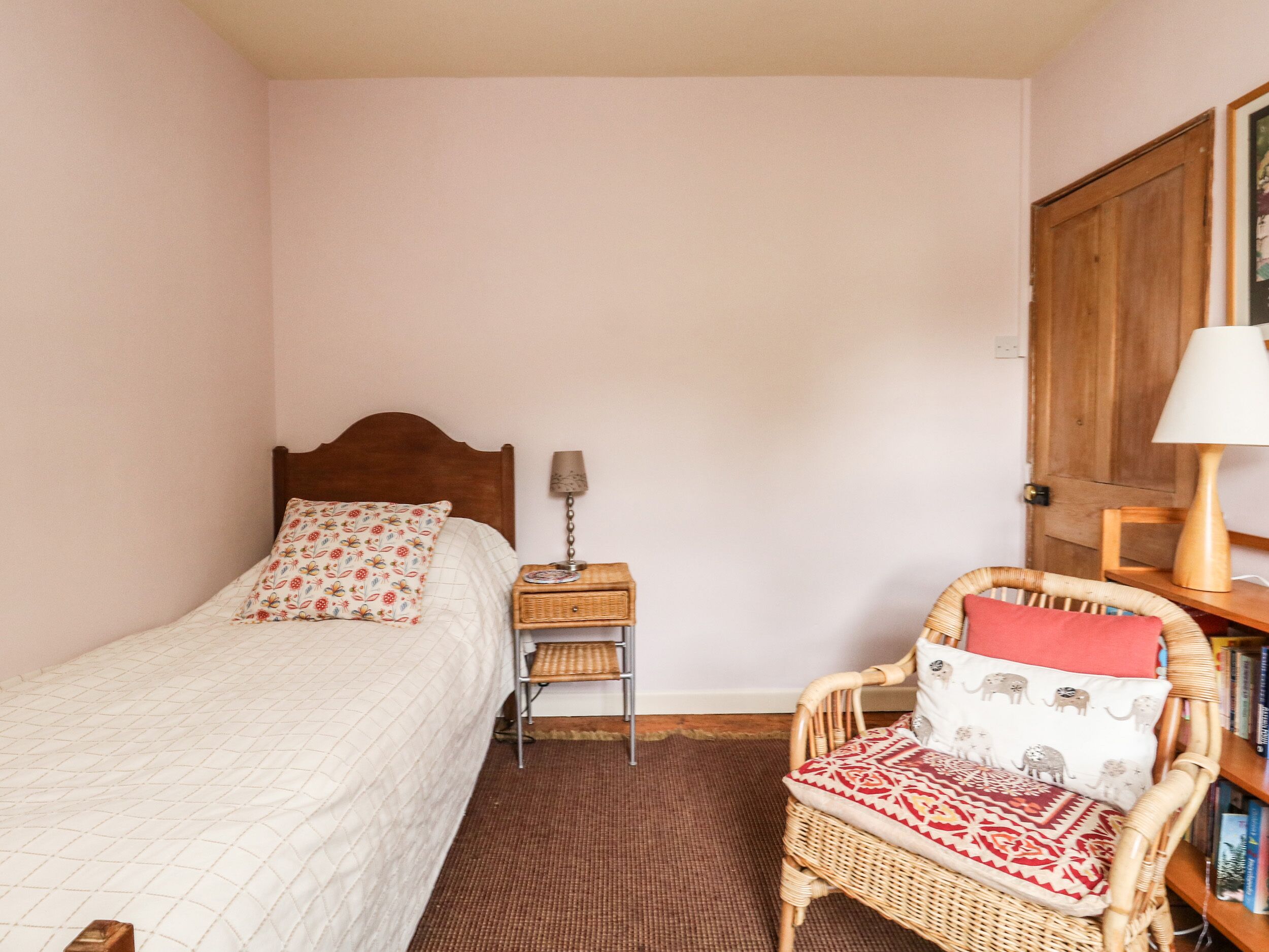 A bedroom with a single bed, a nightstand with a lamp, a wicker chair with a cushion, and a bookshelf.