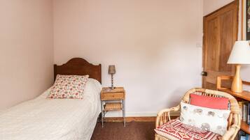 A bedroom with a single bed, a nightstand with a lamp, a wicker chair with a cushion, and a bookshelf.