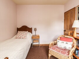 A bedroom with a single bed, a nightstand with a lamp, a wicker chair with a cushion, and a bookshelf.