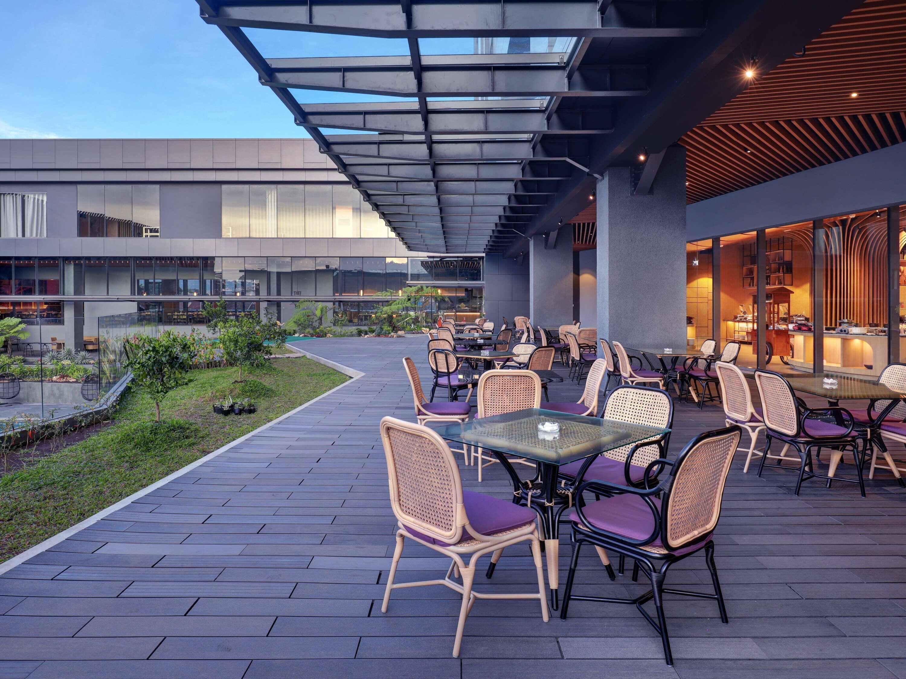 Un patio al aire libre con mesas y sillas, una piscina y un edificio con grandes ventanales al fondo.