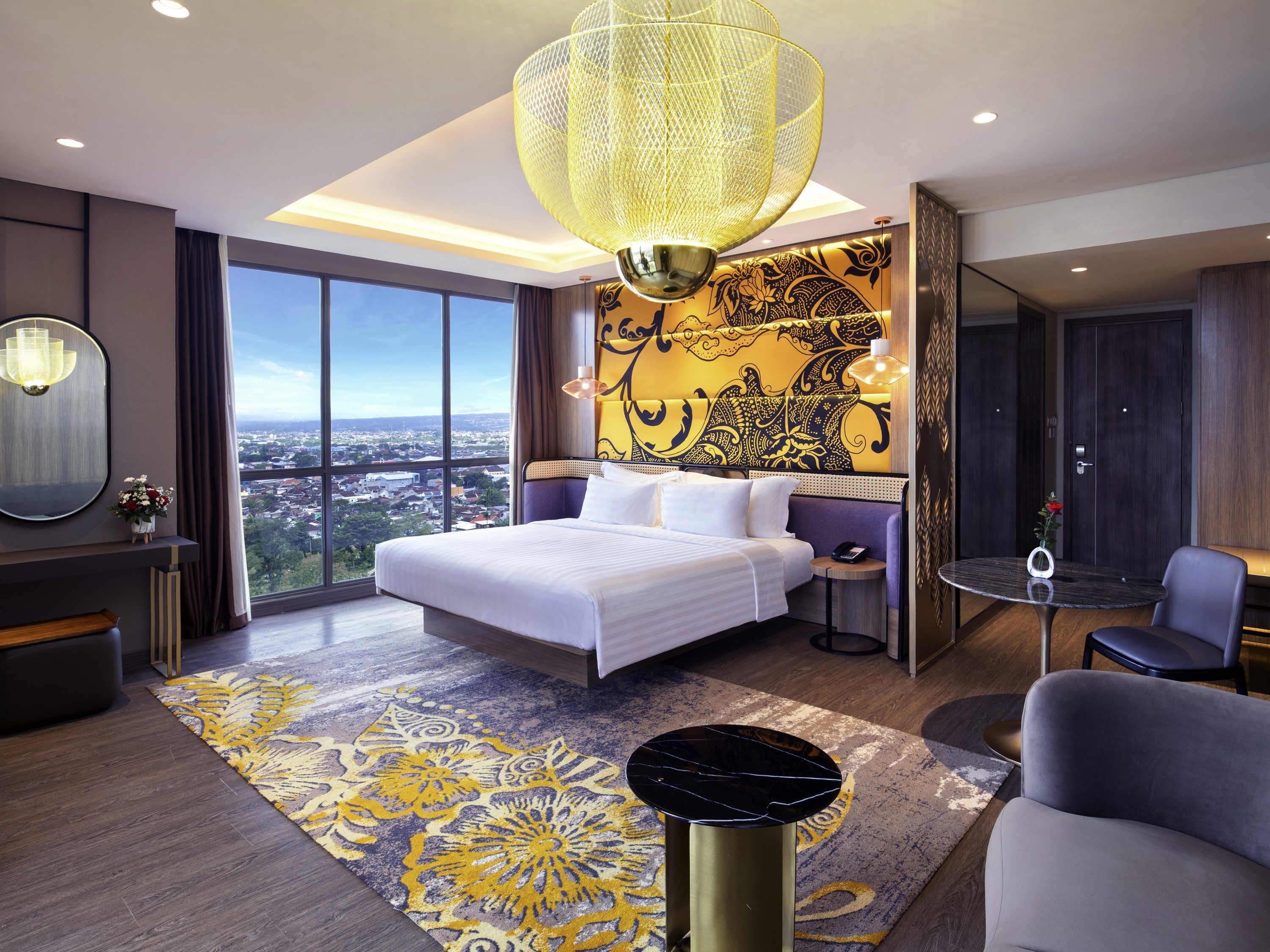 A hotel room with a large bed, a view of the city, a decorative wall, and a modern chandelier.