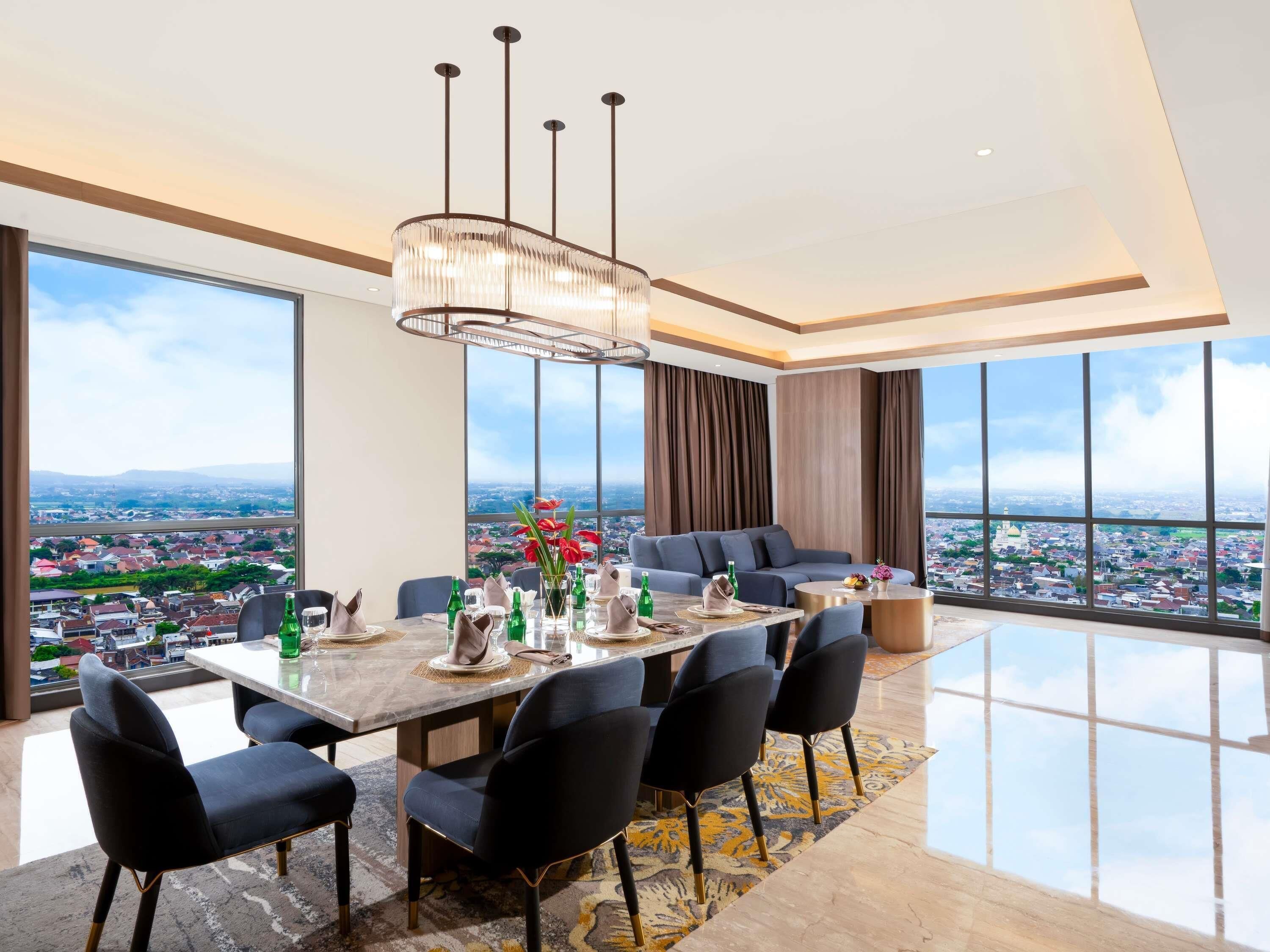 A modern dining area with a large table, chairs, and a chandelier. There is a sofa and a large window with a city view.