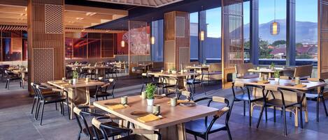 A spacious restaurant with multiple tables and chairs, large windows offering a view of the cityscape, and a modern interior design.