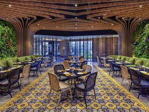 Um restaurante com interior moderno, apresentando uma área de jantar central com mesas e cadeiras redondas, rodeada por assentos estofados verdes.