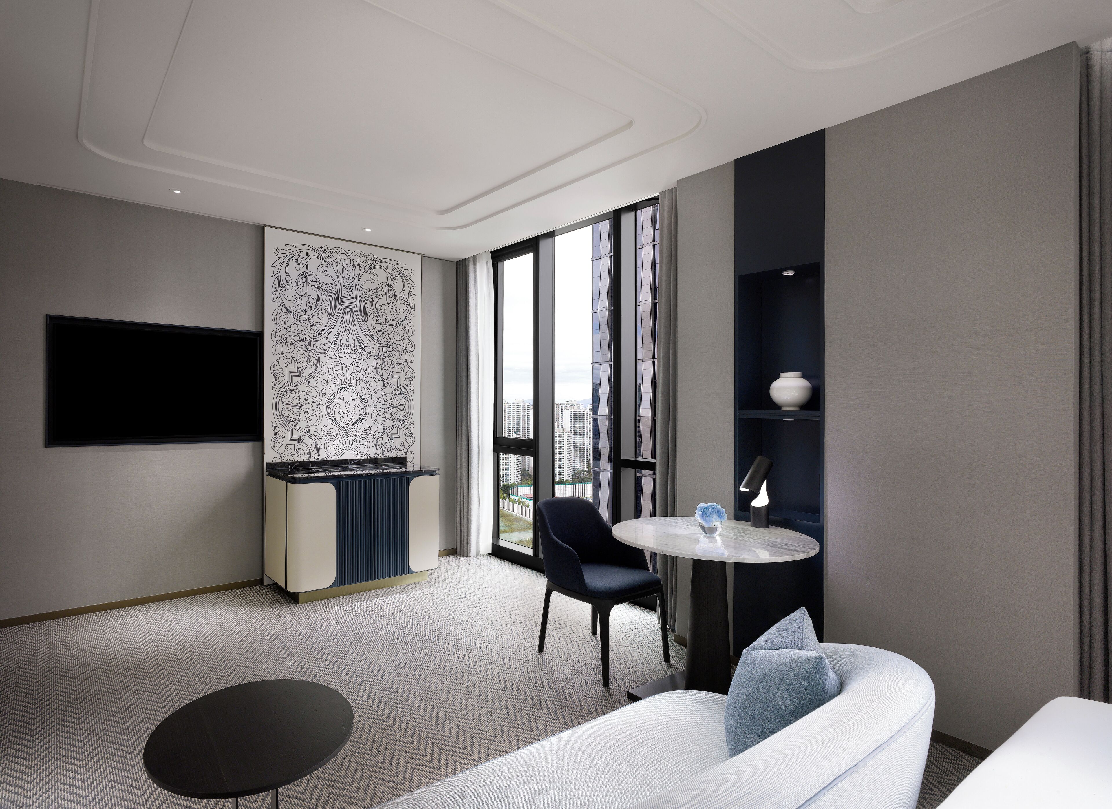 A modern hotel room with a large window, a flat-screen TV, a sofa, a small round table, and a decorative wall panel.