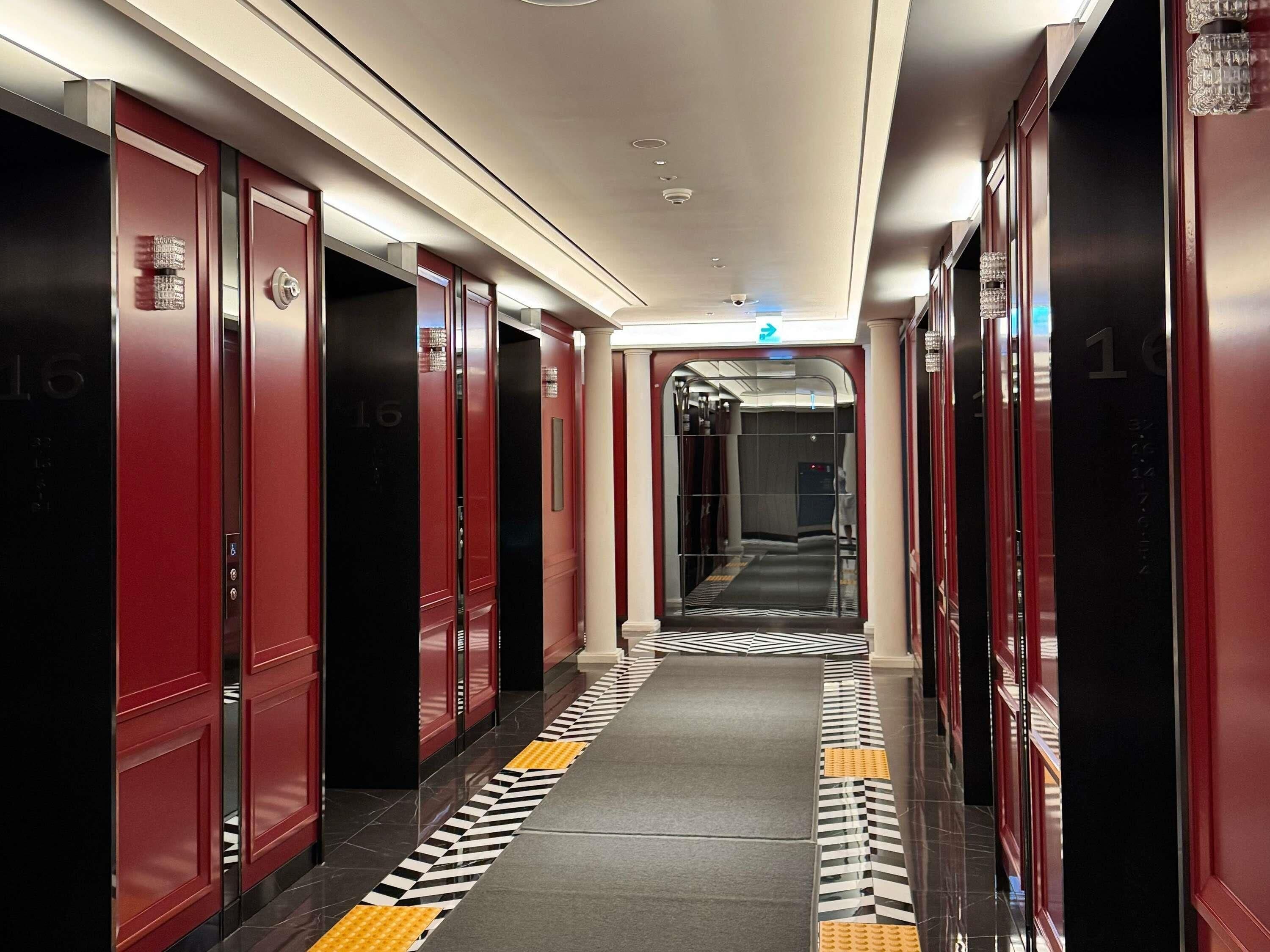 Pasillo de hotel con puertas rojas, piso de estampado blanco y negro y un ascensor central.