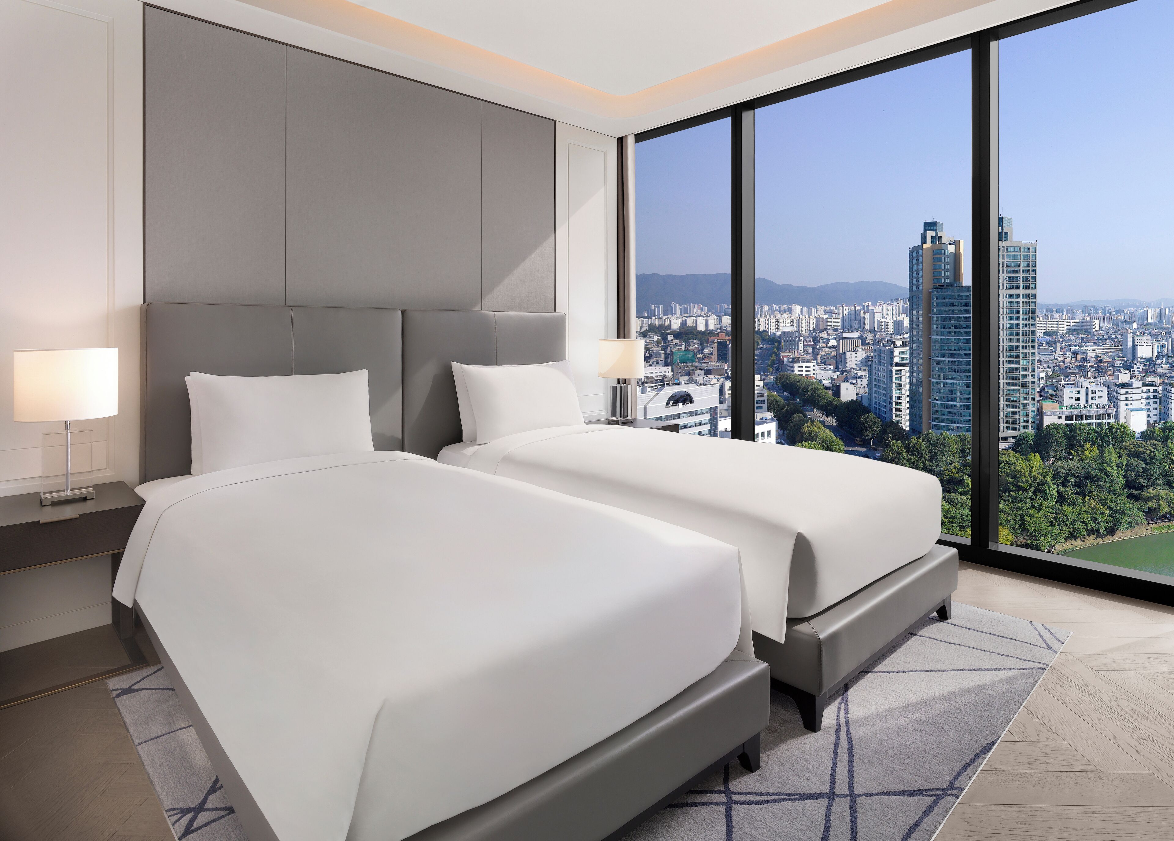 A modern hotel room with two beds, a large window with a city view, and a grey headboard.
