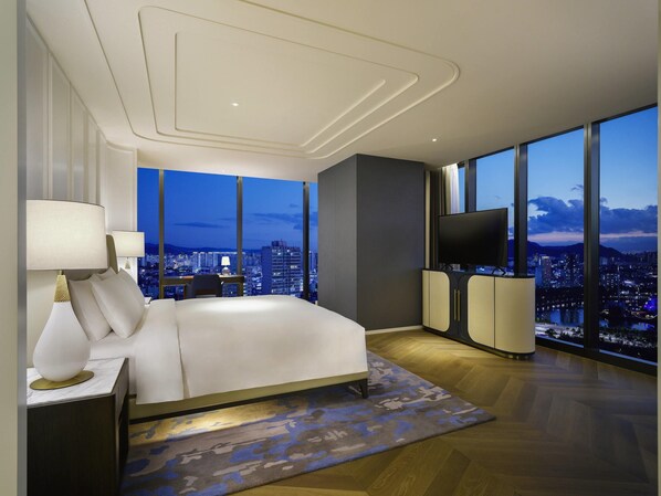 A modern bedroom with a city view, a large bed, a TV, and a desk with a lamp.