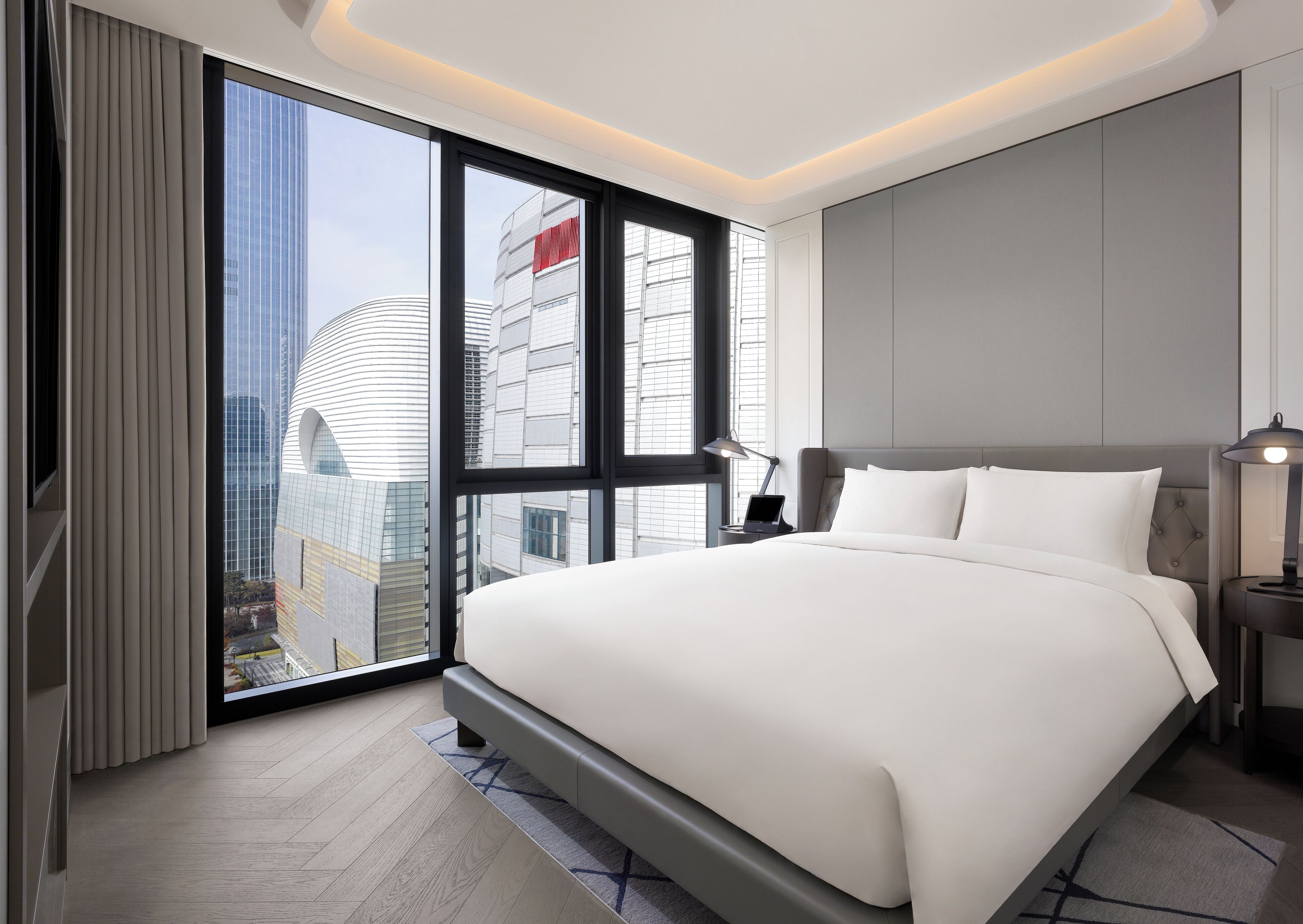A modern hotel room with a large bed, a desk with a laptop, a nightstand with a lamp, and a view of a cityscape through floor-to-ceiling windows.