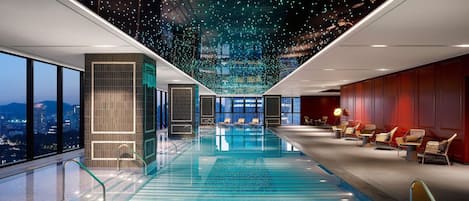 A modern indoor swimming pool with a glass floor, surrounded by seating areas and large windows offering city views.