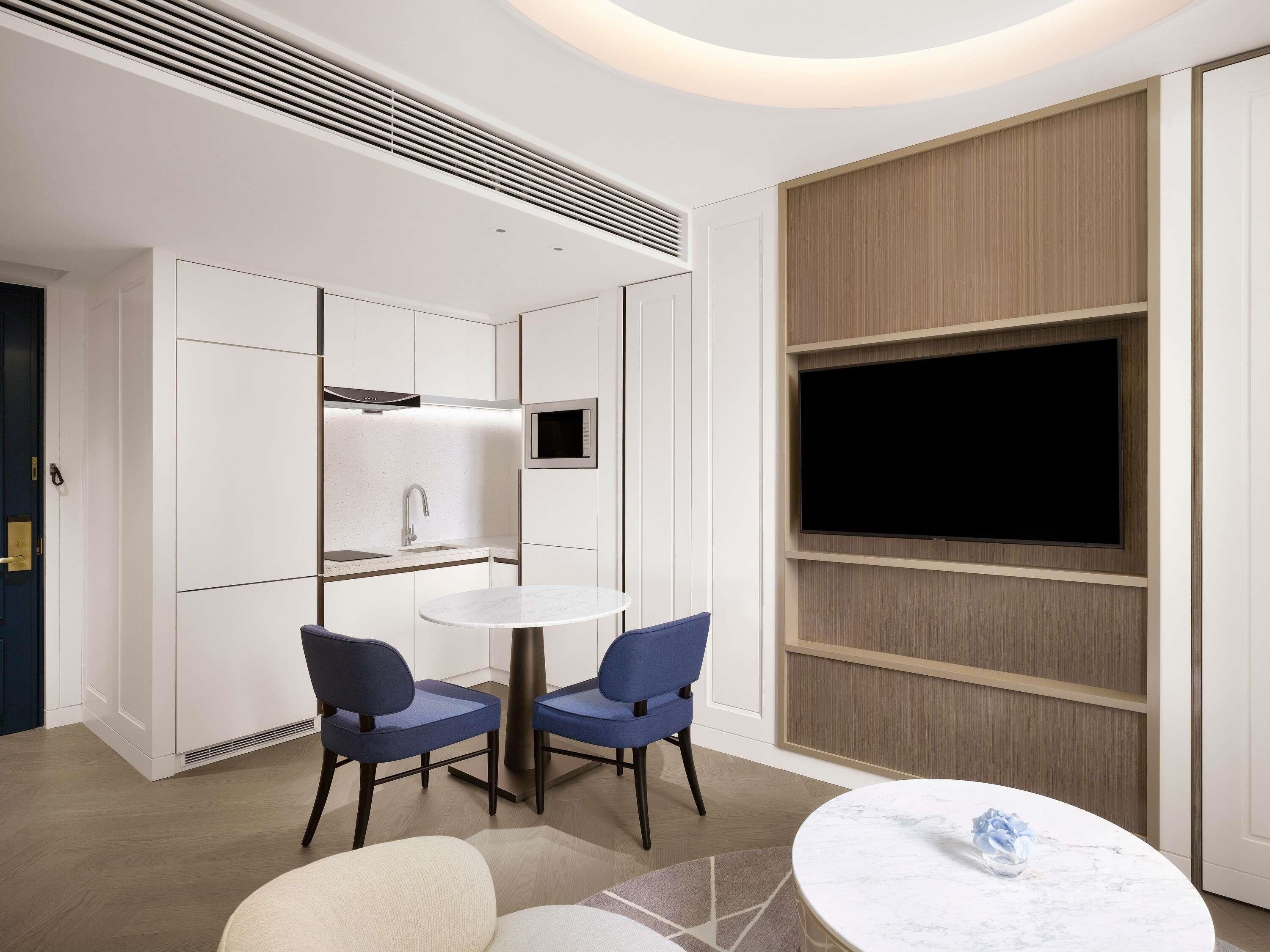 A modern hotel room with a kitchenette, a round dining table, two chairs, a TV mounted on the wall, and a sofa.