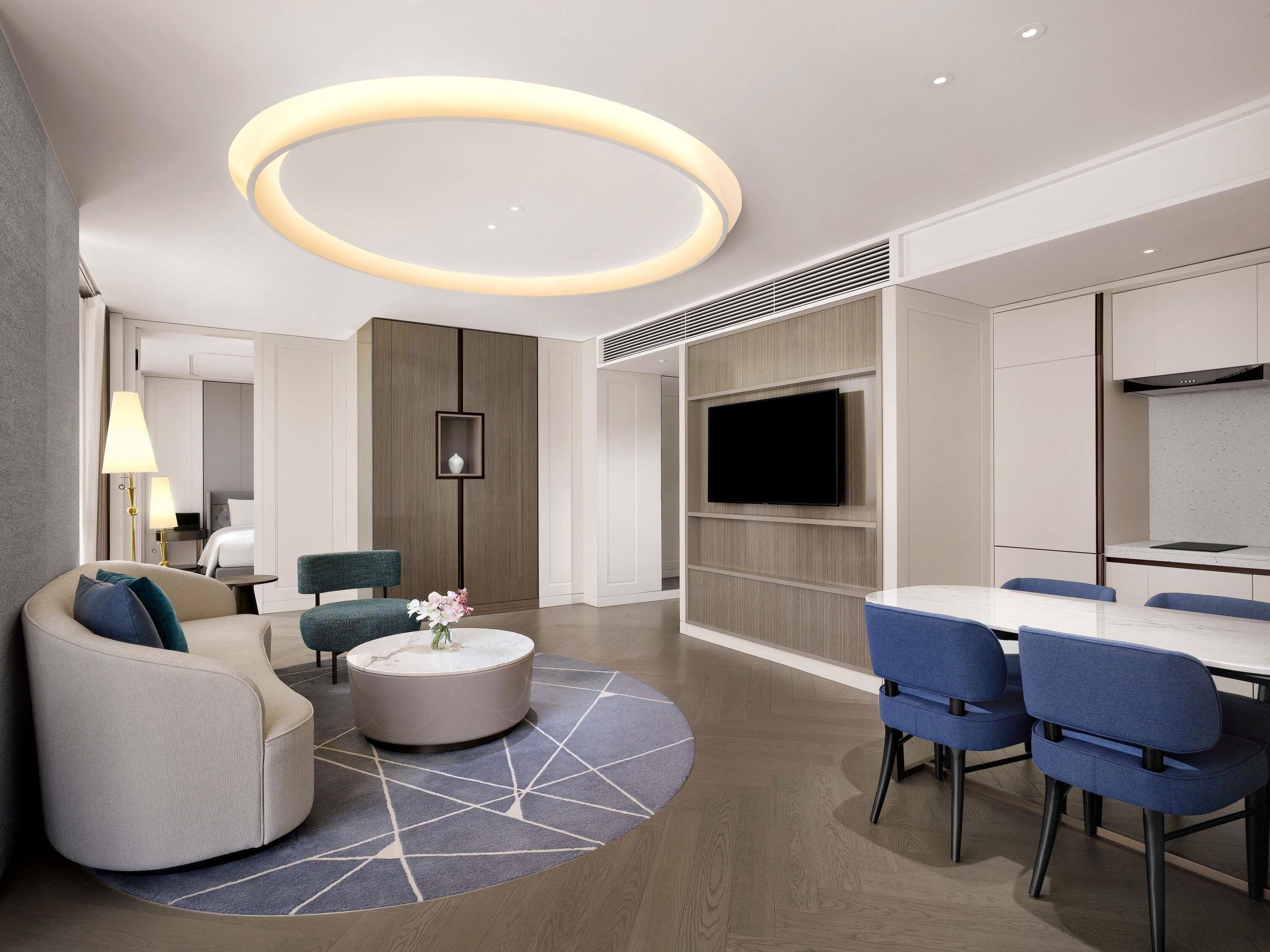 Una habitación de hotel moderna con una lámpara de techo redonda, un televisor de pantalla plana, un comedor con sillas azules y un sofá con un tapete de diseño geométrico.