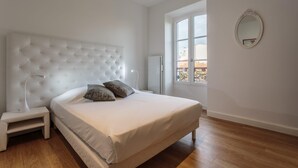 Une chambre à coucher avec une tête de lit capitonnée, une table de chevet, une commode, un lit avec deux oreillers, un miroir et une fenêtre donnant sur un beau paysage.