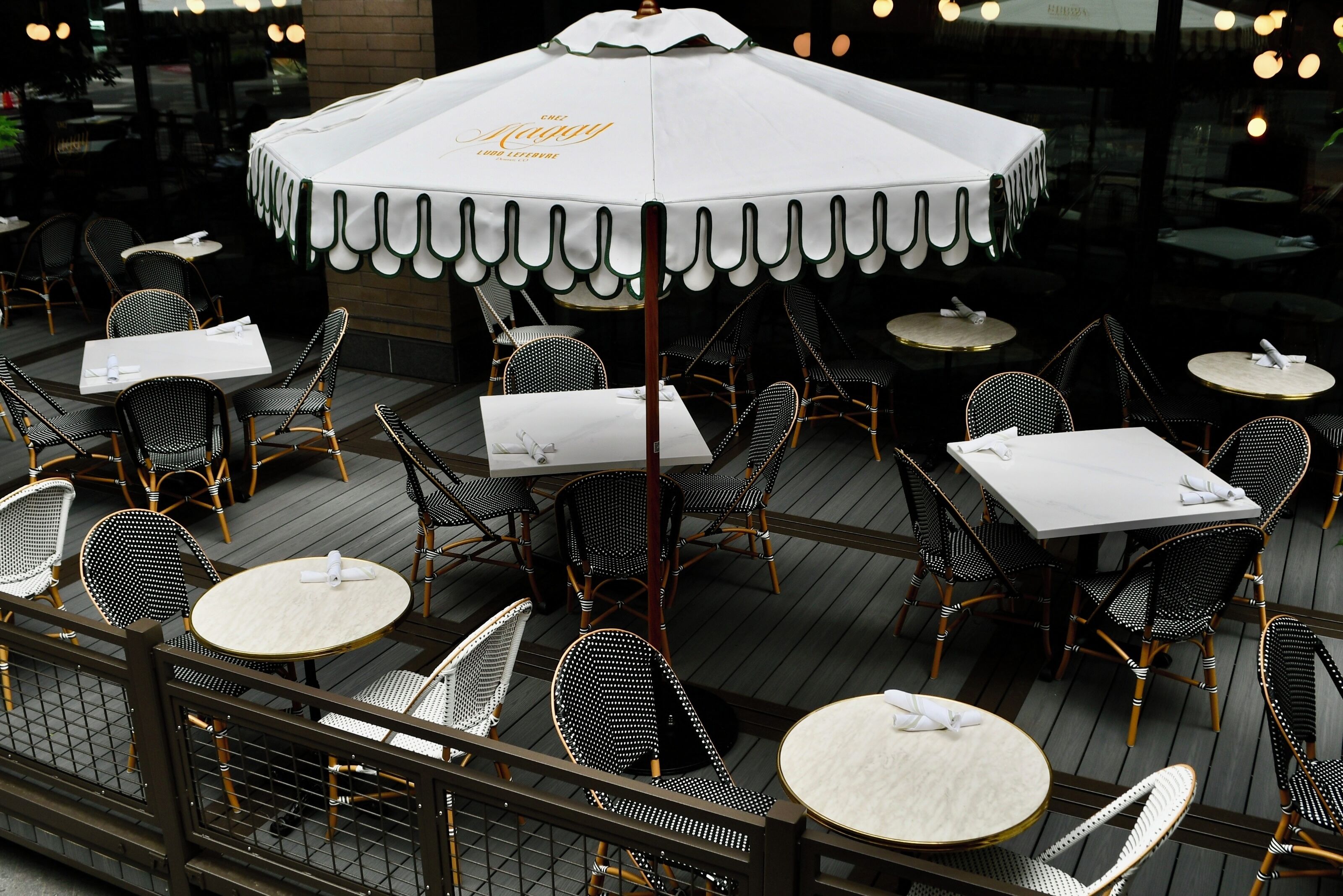 An outdoor dining area with round tables and black wicker chairs, covered by a white umbrella with a scalloped edge.
