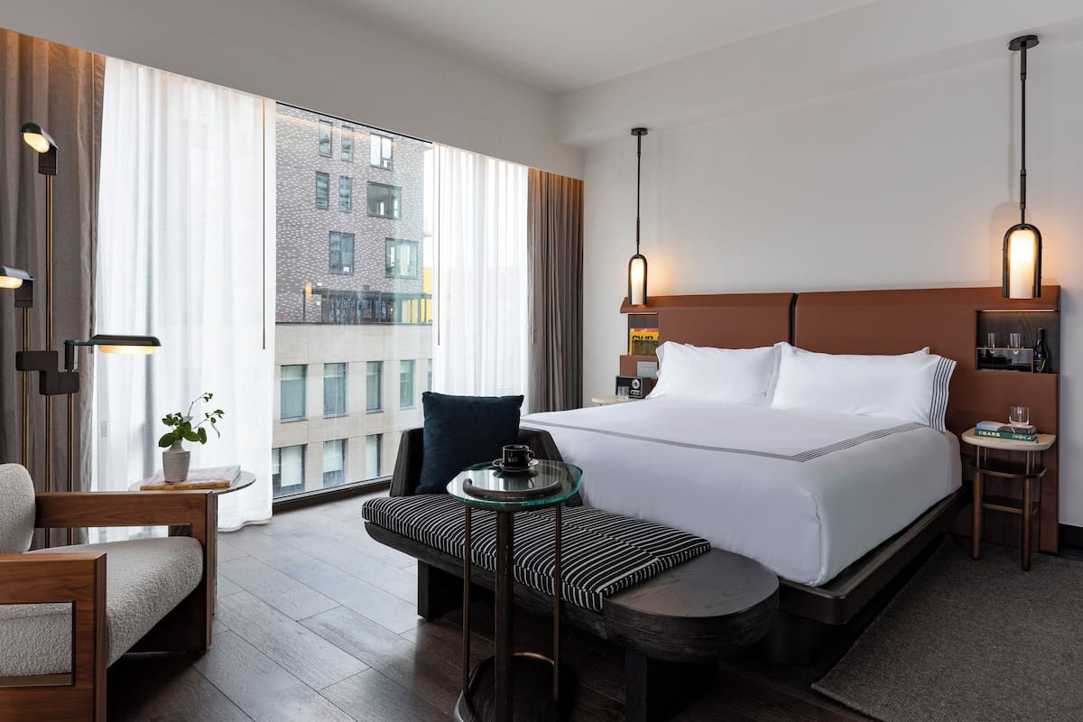 Ein modernes Hotelzimmer mit einem großen Bett, einer Sitzecke mit kleinem Tisch und Blick auf eine Stadtlandschaft durch große Fenster.