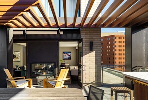 Un área de estar moderna al aire libre con mobiliario de madera, cerramiento de vidrio y vista a un paisaje urbano.