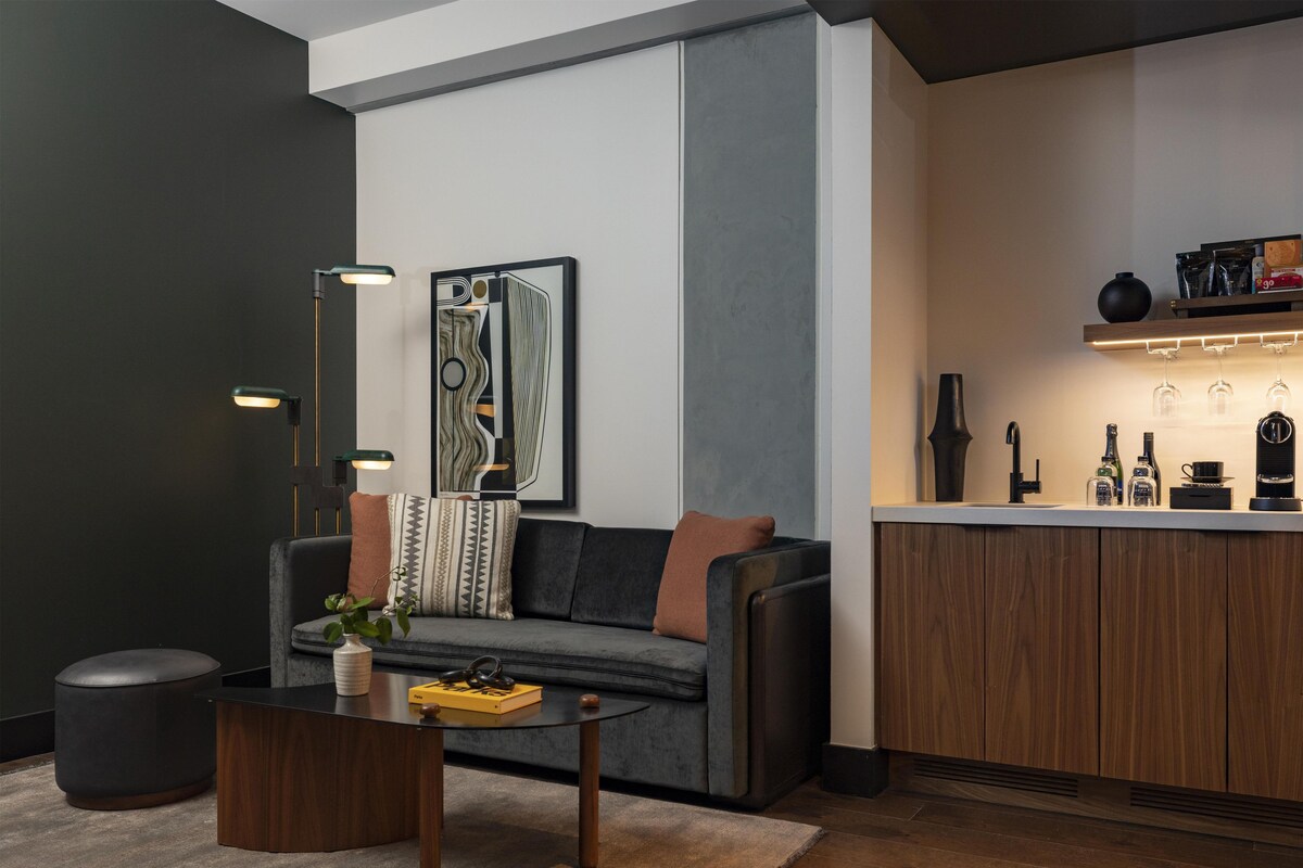 A modern living room with a dark grey sofa, a coffee table, and a kitchen area with a wooden cabinet and a shelf with bottles.