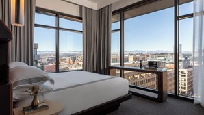 Ein modernes Hotelzimmer mit einem großen Bett, einem Nachttisch mit Buch und Vase sowie Blick auf die Stadt durch große Fenster.