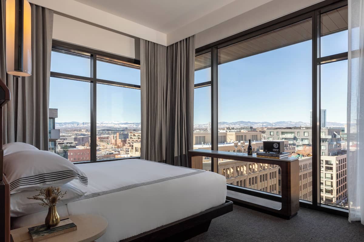 Ein modernes Hotelzimmer mit einem großen Bett, einem Nachttisch mit Buch und Vase sowie Blick auf die Stadt durch große Fenster.