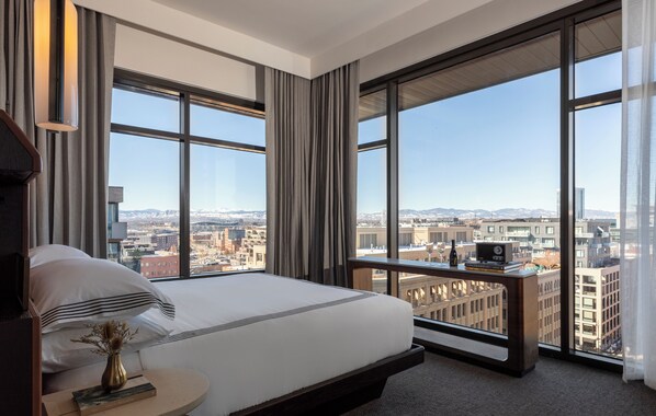 Habitación de hotel moderna con una cama grande, una mesita de noche con un libro y un florero, y vistas de la ciudad a través de amplias ventanas.