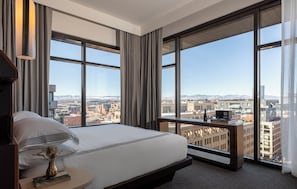 A modern hotel room with a large bed, a bedside table with a book and a vase, and a view of the city through large windows.