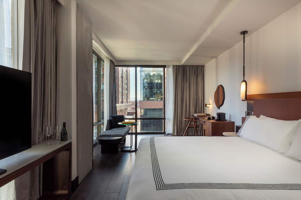 Ein Hotelzimmer mit einem großen Bett, einem Fernseher, einem Schreibtisch mit Stuhl und Blick auf Gebäude durch das Fenster.
