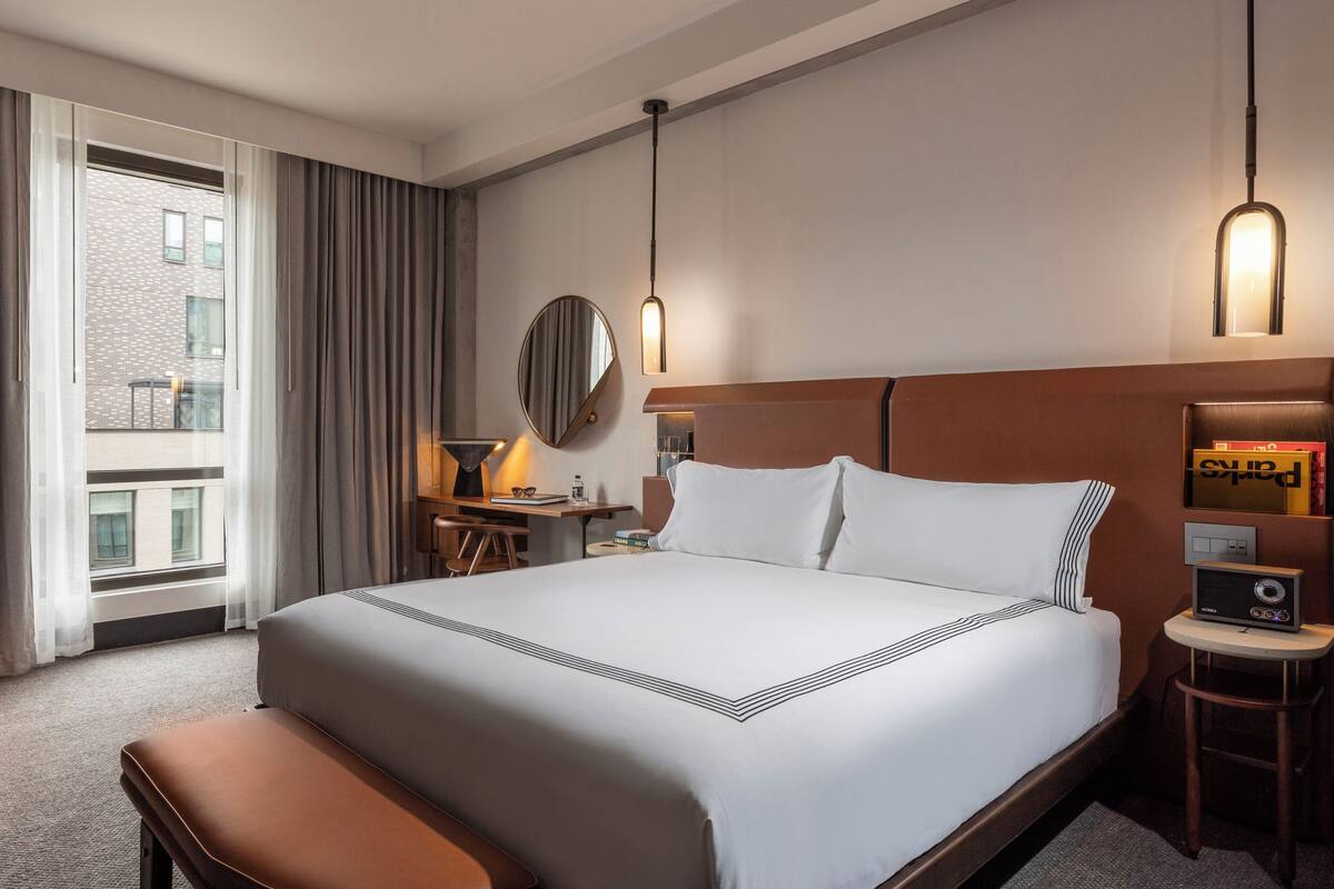 Ein Hotelzimmer mit einem großen Bett, einem Schreibtisch mit Lampe, einem runden Spiegel und Blick auf ein Gebäude durch das Fenster.