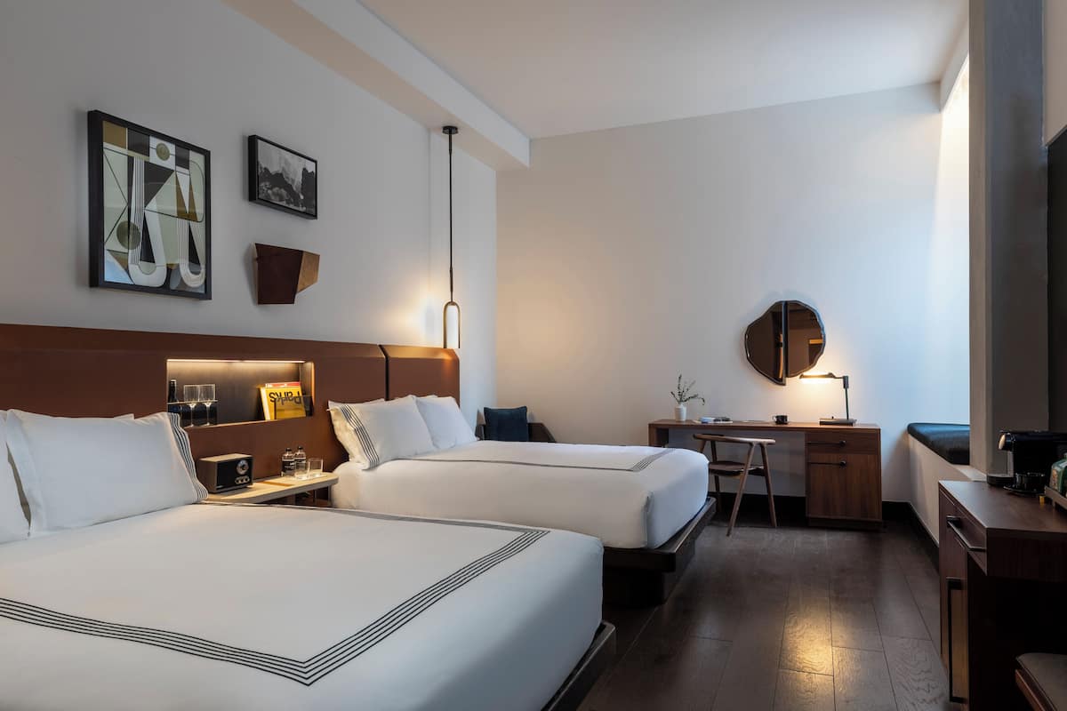 Ein Hotelzimmer mit zwei Betten, einem Schreibtisch, einem Stuhl, einem kleinen Tisch, einem Spiegel und Bildern an der Wand.