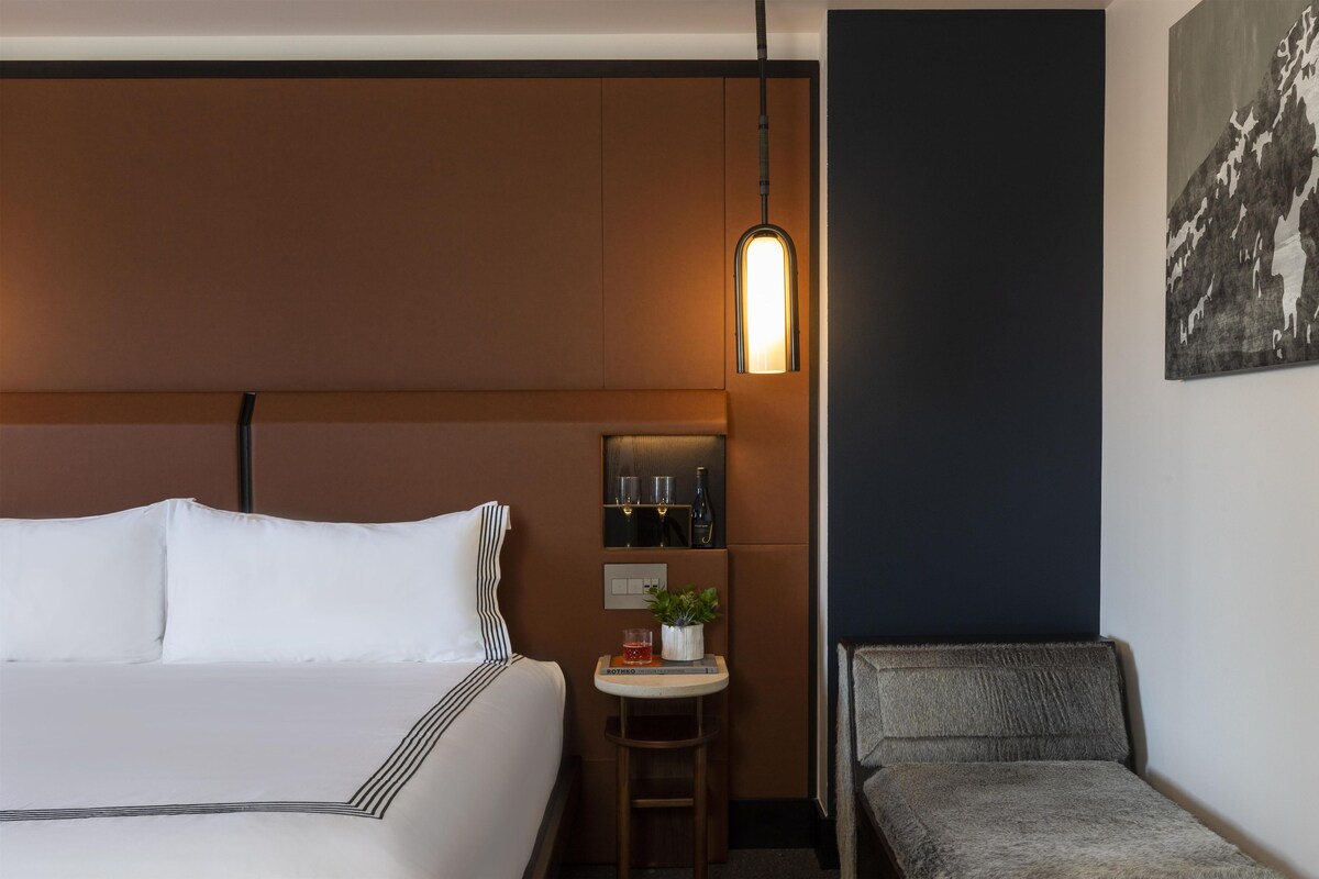 A hotel room with a bed, a small table with a plant, a chair, and a wall-mounted shelf with glasses and bottles.
