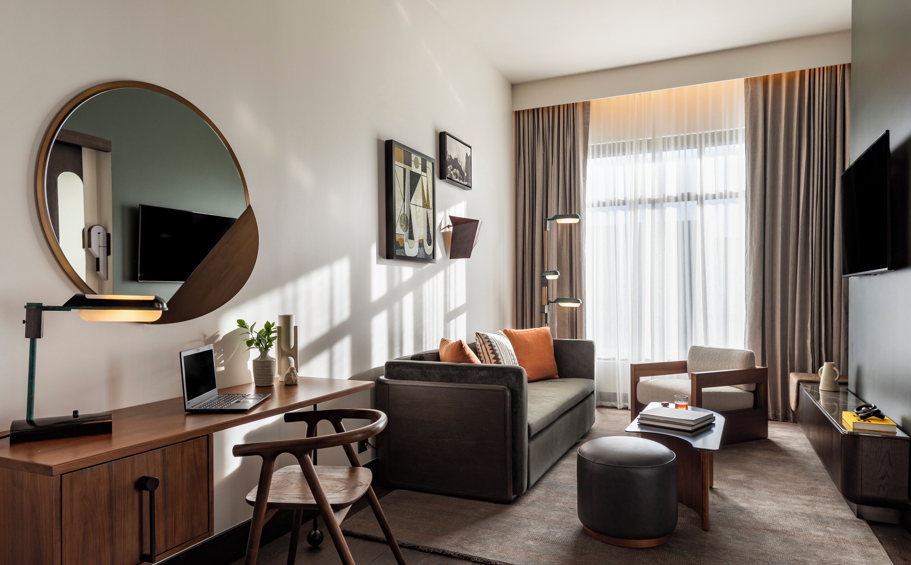 A modern living room with a sofa, a coffee table, a desk with a laptop, a chair, a round ottoman, and a large mirror.