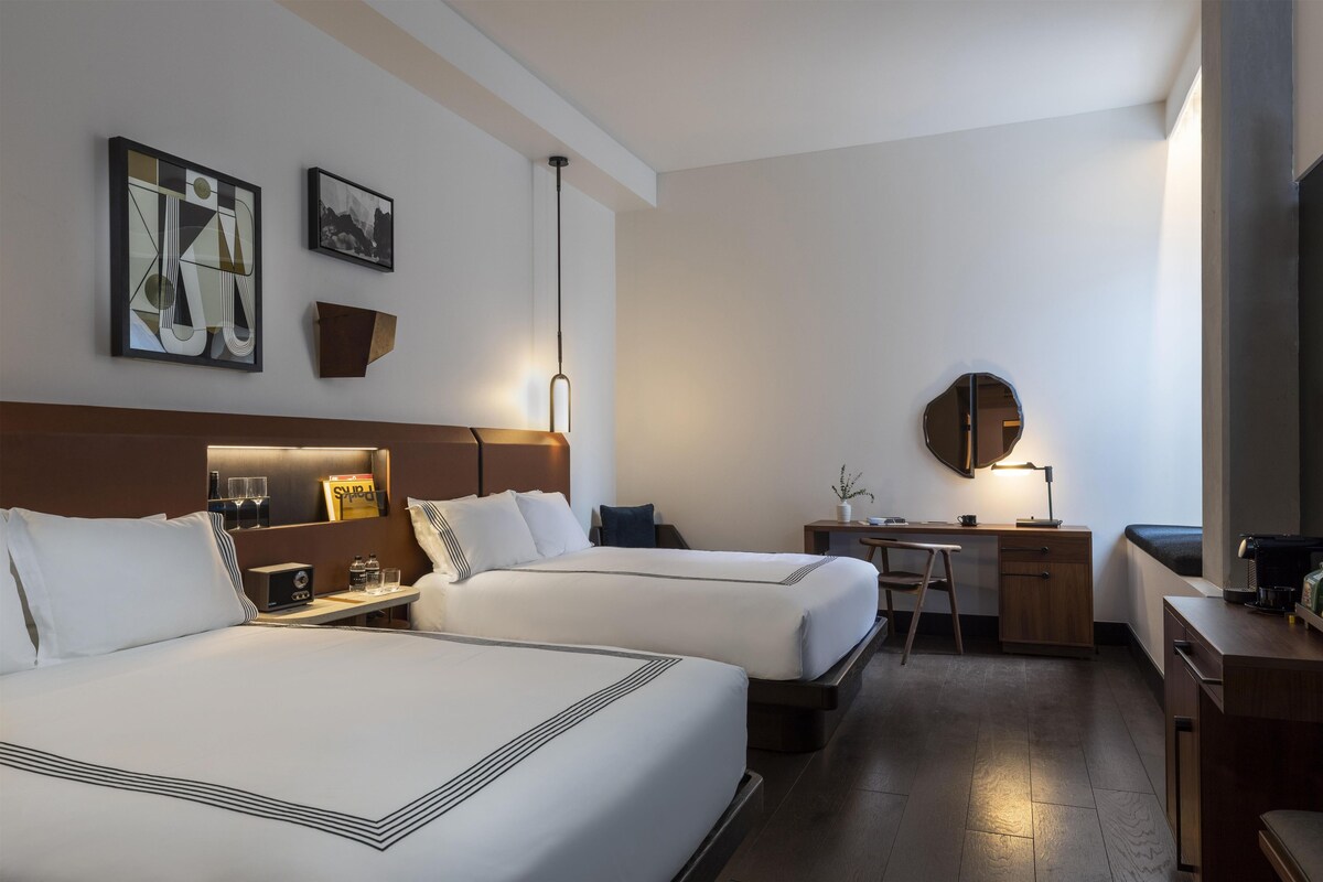 A hotel room with two beds, a desk, a chair, a coffee machine, and a television.