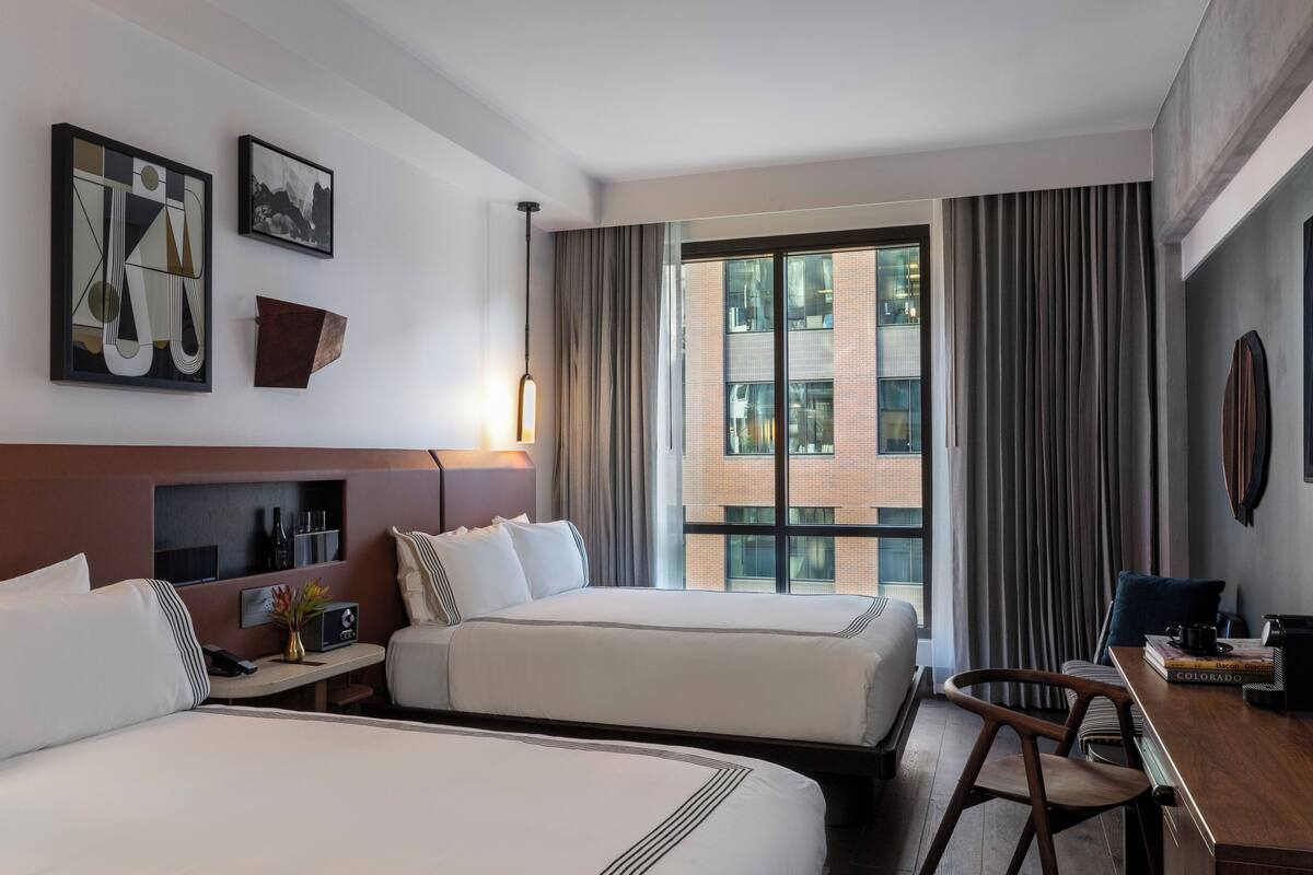 Ein Hotelzimmer mit zwei Betten, einem Schreibtisch, einem Stuhl, einem Fenster mit Vorhängen und Bildern an den Wänden.