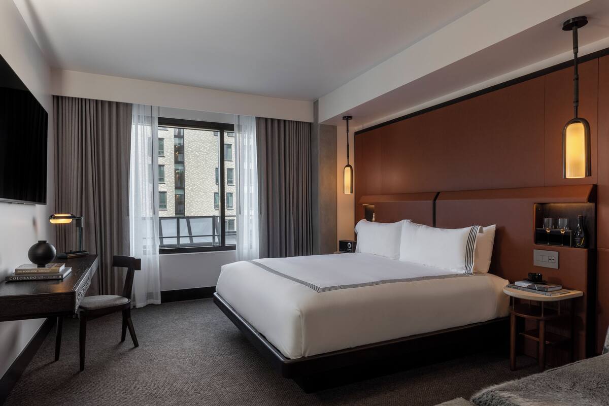 Ein Hotelzimmer mit einem großen Bett, einem Schreibtisch mit Lampe, einem Sessel, einem kleinen Tisch und einem Fenster mit Vorhängen.