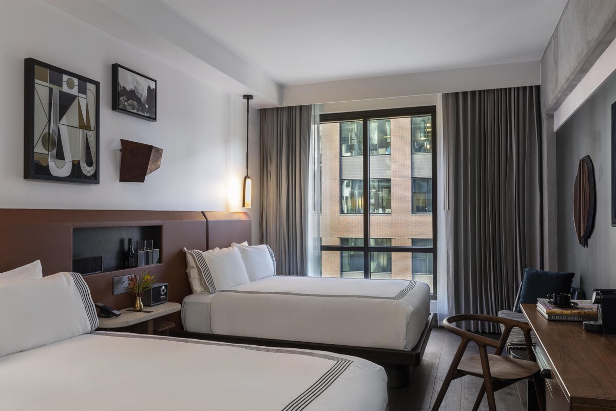 A hotel room with two beds, a desk, a chair, a window with curtains, and artwork on the wall.