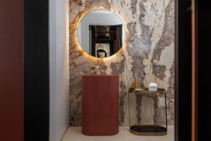A bathroom with a marble wall, a round mirror, a red sink, and a small shelf with towels.