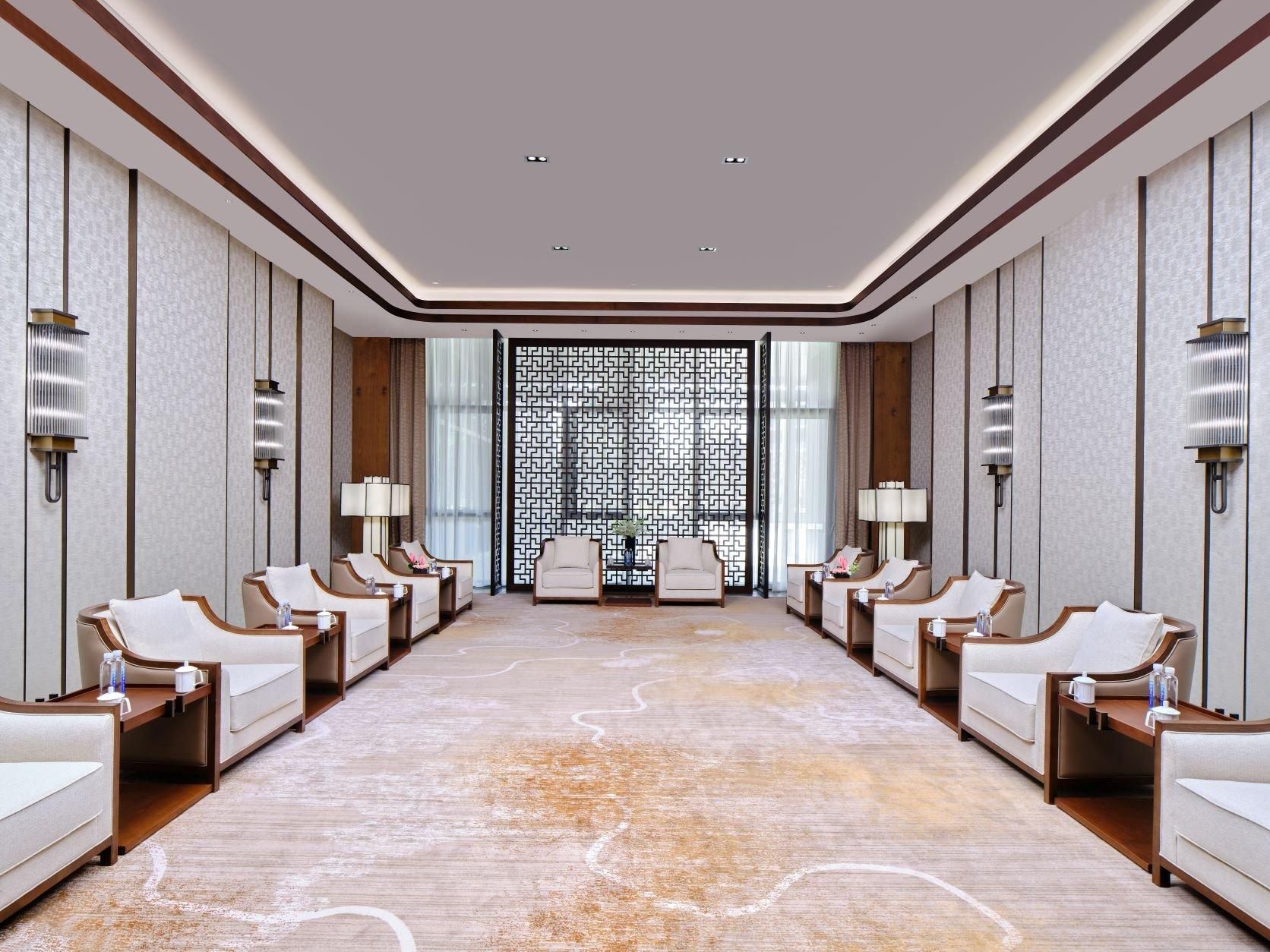 A hotel room with a large, patterned carpet, a central seating area with chairs and tables, and a decorative wall panel.